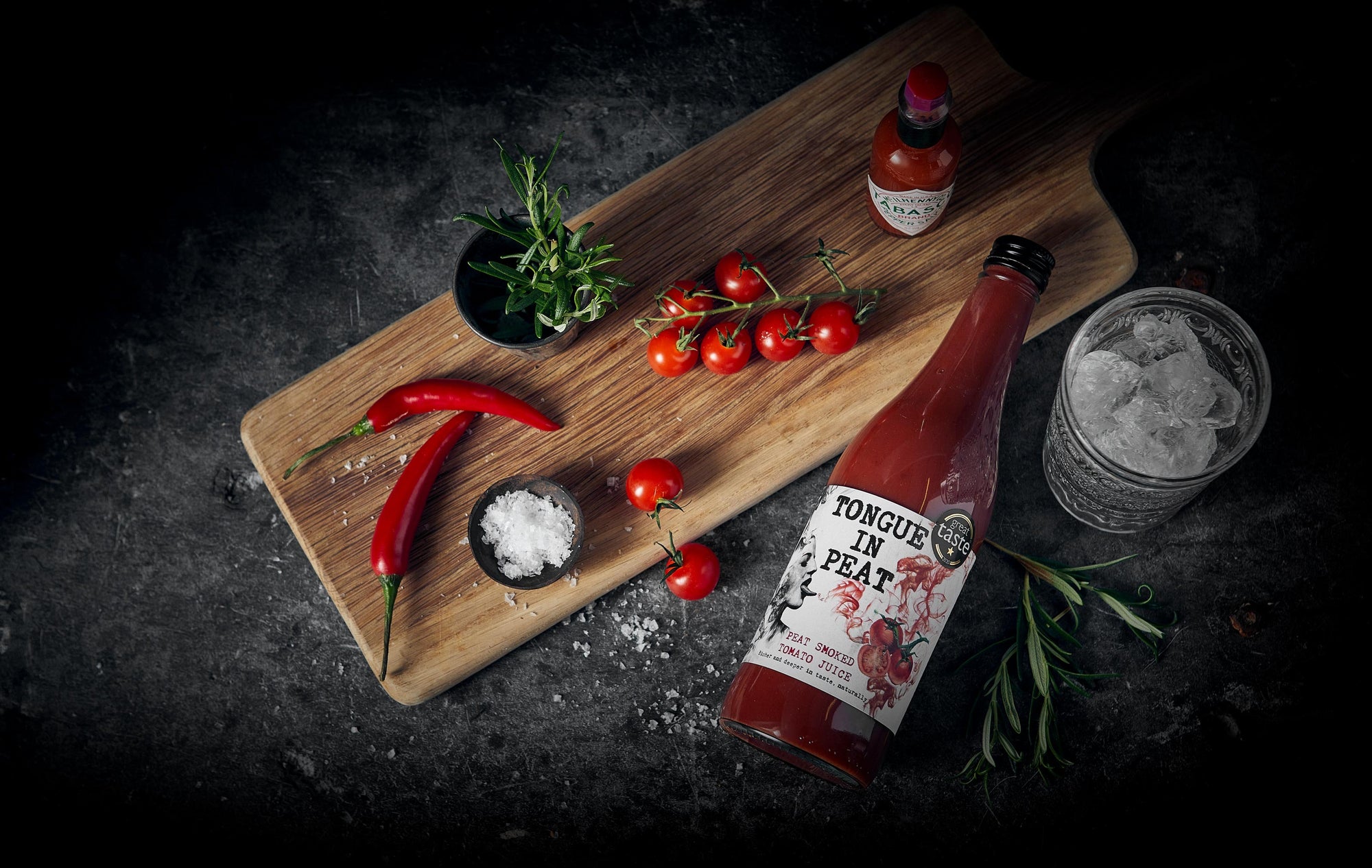 wooden board with chillis, rosemary, cherry tomatoes and tabasco on top next to a glass of ice and a bottle of peat smoked tomato juice by Tongue in Peat
