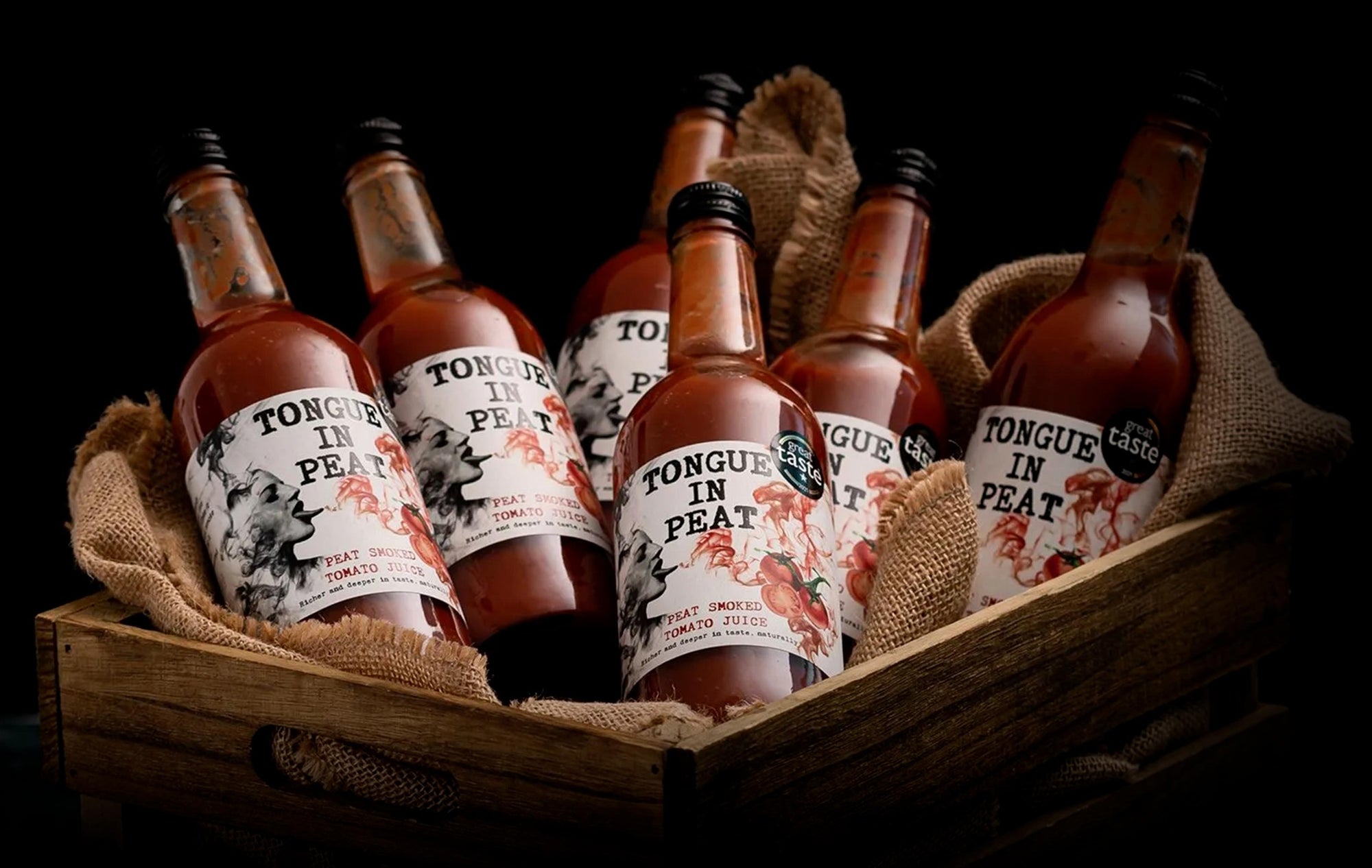 6 peat smoked tomato juice bottles by Tongue in Peat lying on top of a cloth inside a wooden basket