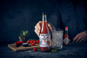 Tongue in Peat smoked tomato juice on a counter next to tabasco, chilli, sea salt, cherry tomatoes, with a person standing behind and holding the bottle
