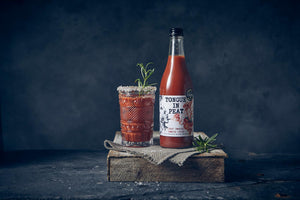 Bloody Mary cocktail garnished with rosemary and a ring of sea salt around the rim next to a bottle of Tongue in Peat smoked tomato juice