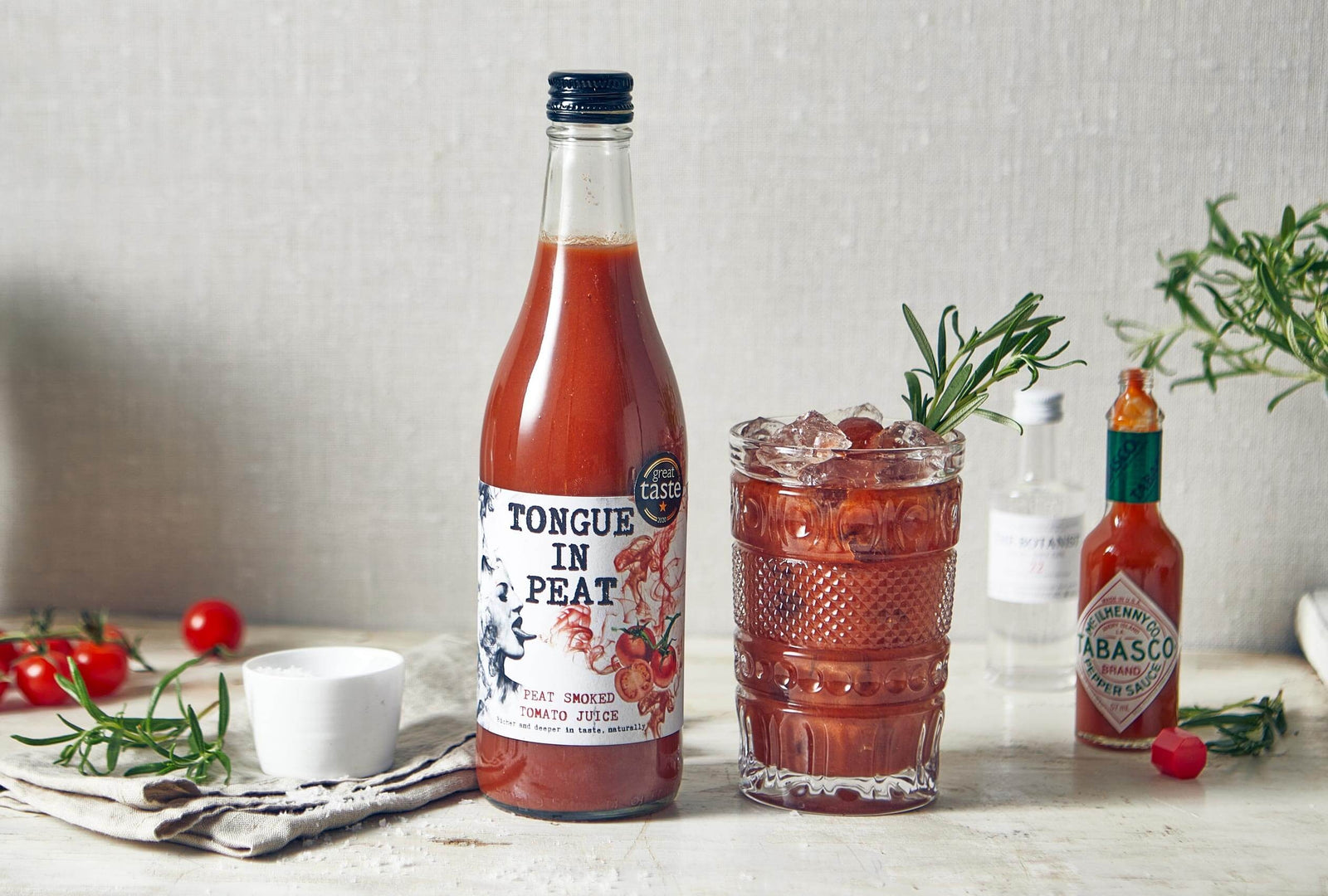 Bottle of peat smoked tomato juice by Tongue in Peat next to a Bloody Mary cocktail garnished with rosemary and two bottles - tabasco and Vodka in the background