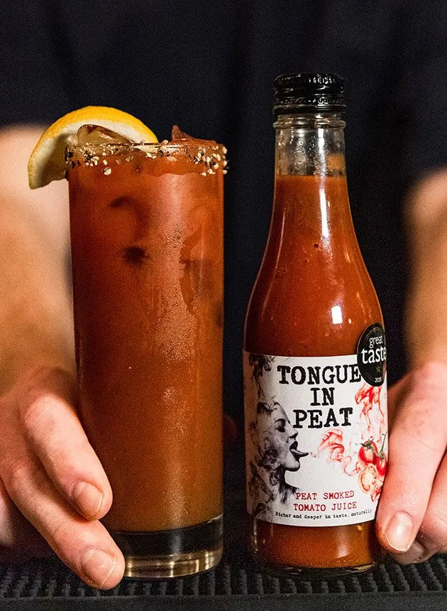 bottle of tongue in peat smoked tomato juice next to a classic Bloody Mary cocktail garnished with a slice of lemon